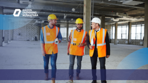 Tiga tenaga profesional konstruksi mengenakan helm dan rompi keselamatan berdiskusi sambil berjalan di dalam area bangunan gedung